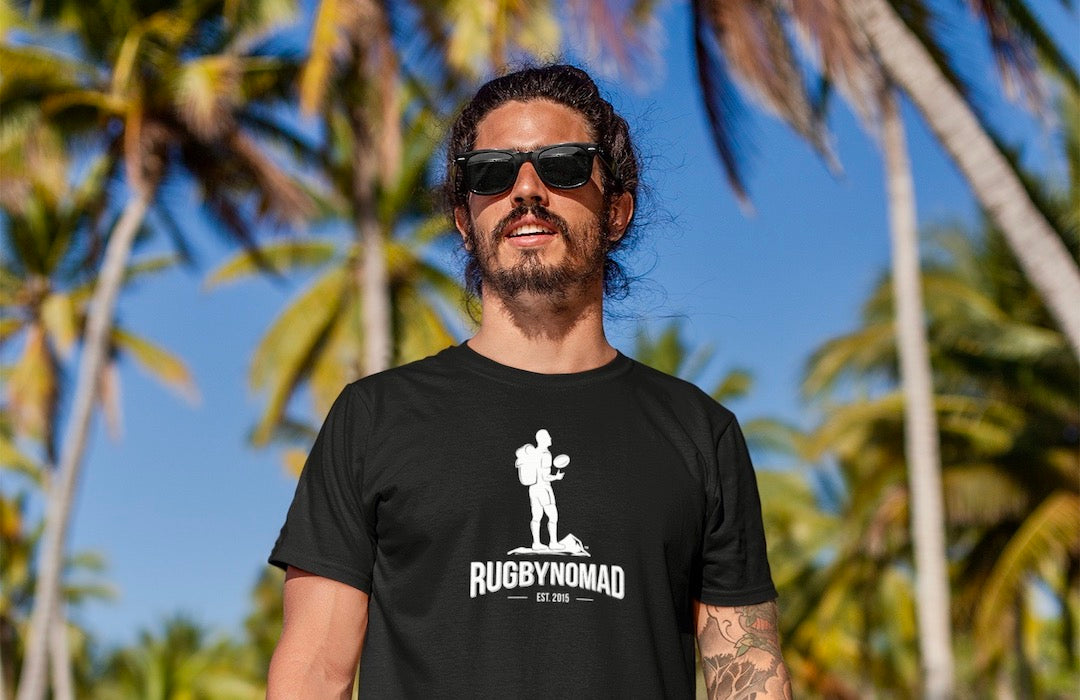 RUGBYNOMAD white color heavyweight eco performance short sleeve t shirt with black original RUGBYNOMAD complete logo on a male model with dark hair and dark beard wearing black sunglasses standing on the beach on sand with green jungle behind him