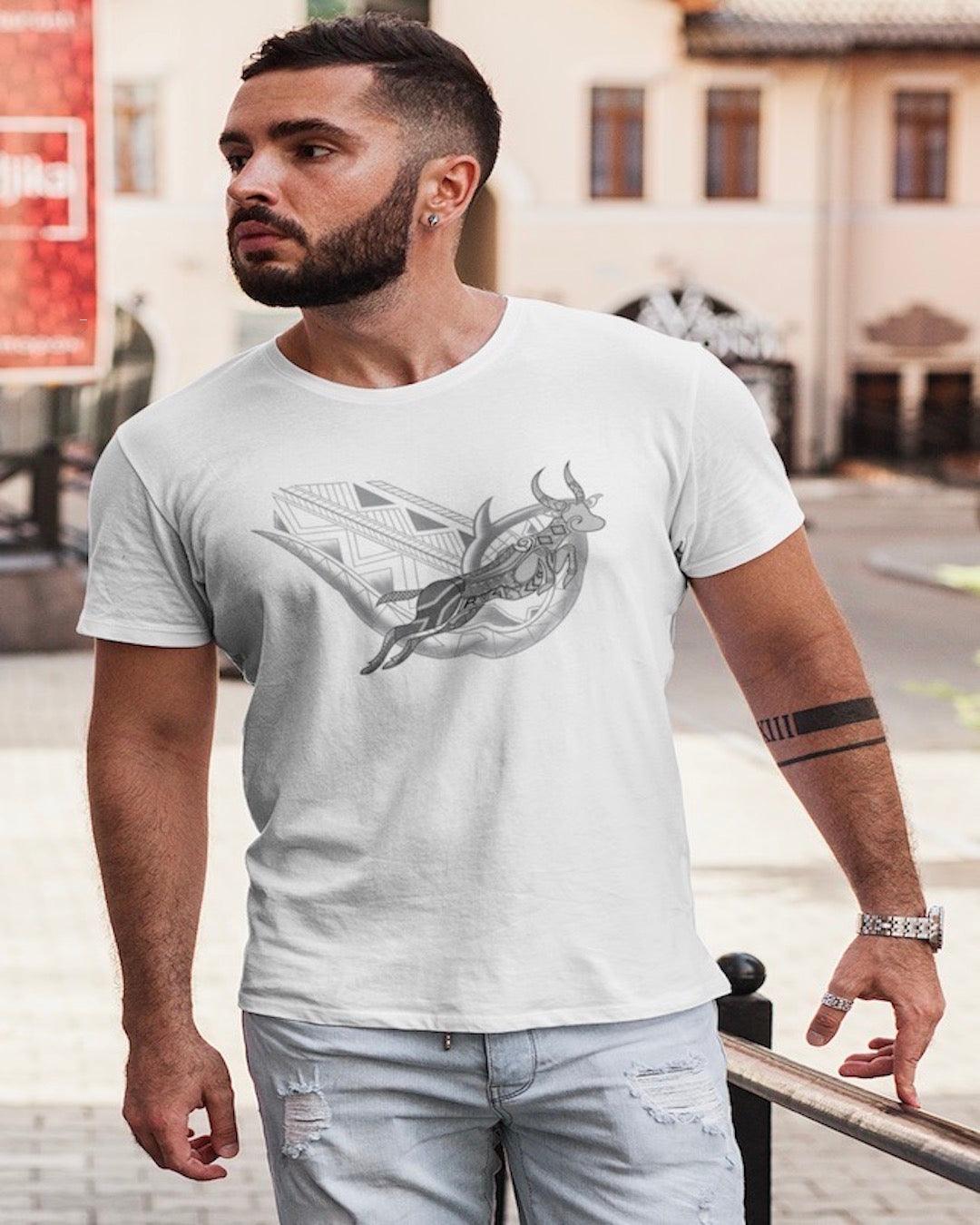 RUGBYNOMAD white organic cotton classic heavyweight streetwear t shirt with a south africa springbok artistic design on a male model with dark hair, dark beard, arm tattoos, wearing light colored blue jeans walking down the street, bright colored buildings behind him Edit