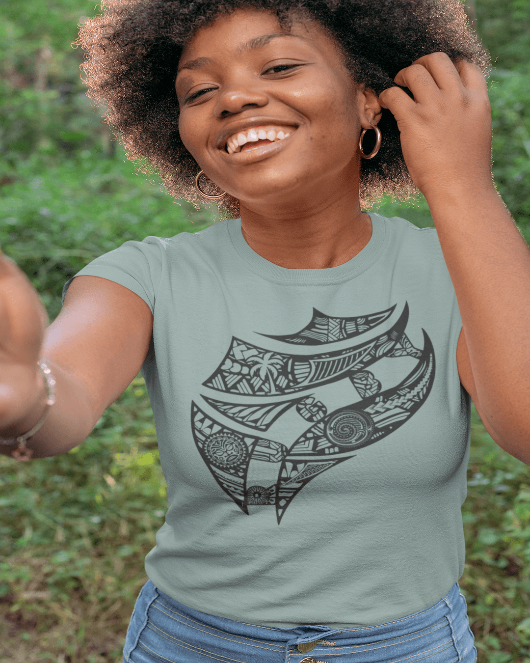 RUGBYNOMAD blue sage color organic cotton women's streetwear t shirt on a female model standing in front of a green jungle with one hand in her dark hair smiling with fiji artistic tribal design