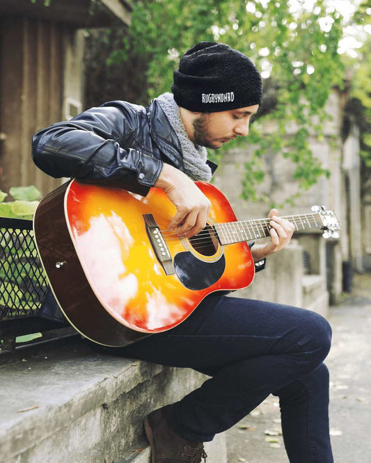 RUGBYNOMAD organic combed cotton black streetwear beanie, male model sitting playing a guitar with RUGBYNOMAD  tribal logo text white