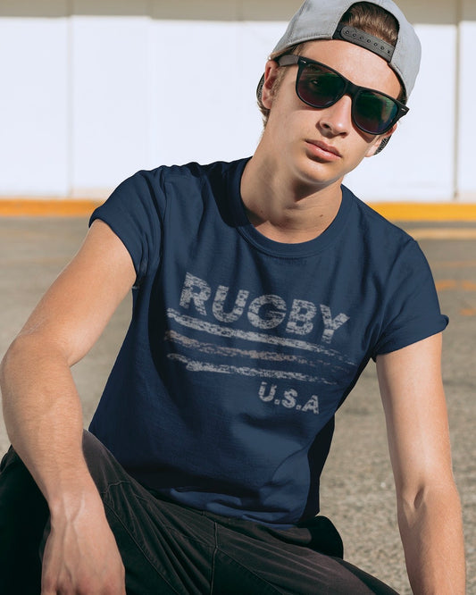 RUGBYNOMAD pacific color organic cotton classic heavyweight streetwear t shirt with a usa rugby artistic design on a male model wearing black pants sitting wearing black sunglasses with a gray baseball cap turned backwards looking straight forward