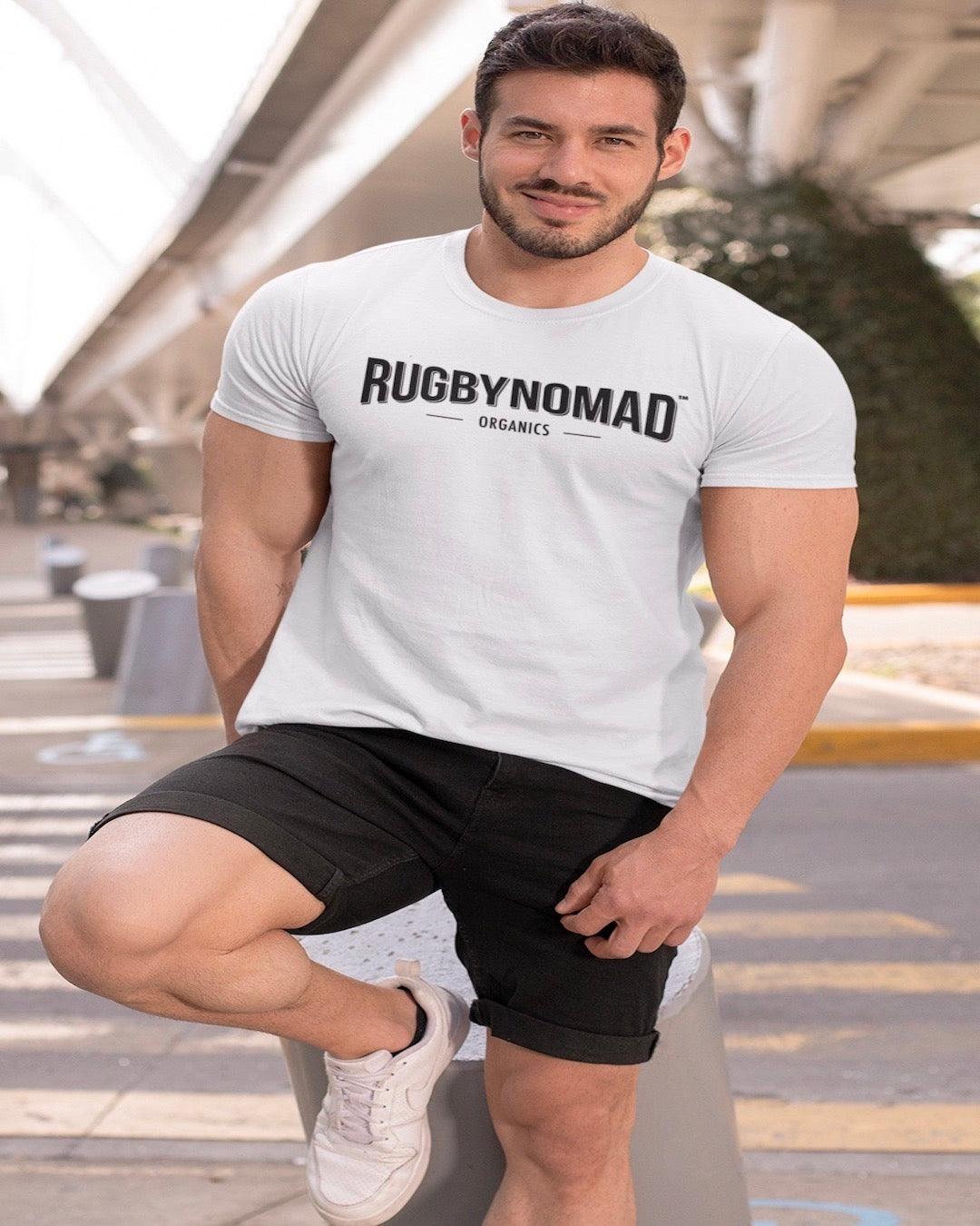 RUGBYNOMAD white color organic cotton heavyweight streetwear t shirt with RUGBYNOMAD original text logo in black on a male model with big muscles wearing black shorts, smiling, black hair, white shoes, leaning on a short post under a bridge