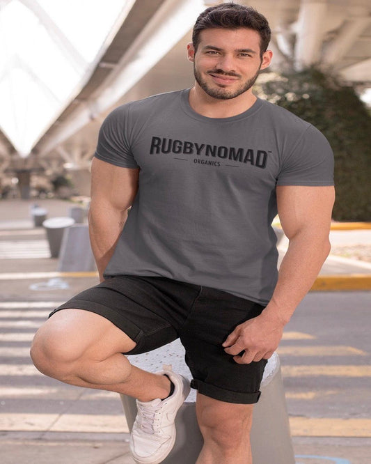 RUGBYNOMAD charcoal color organic cotton heavyweight streetwear t shirt with RUGBYNOMAD original text logo in black on a male model with big muscles wearing black shorts, smiling, black hair, white shoes, leaning on a short post under a bridge