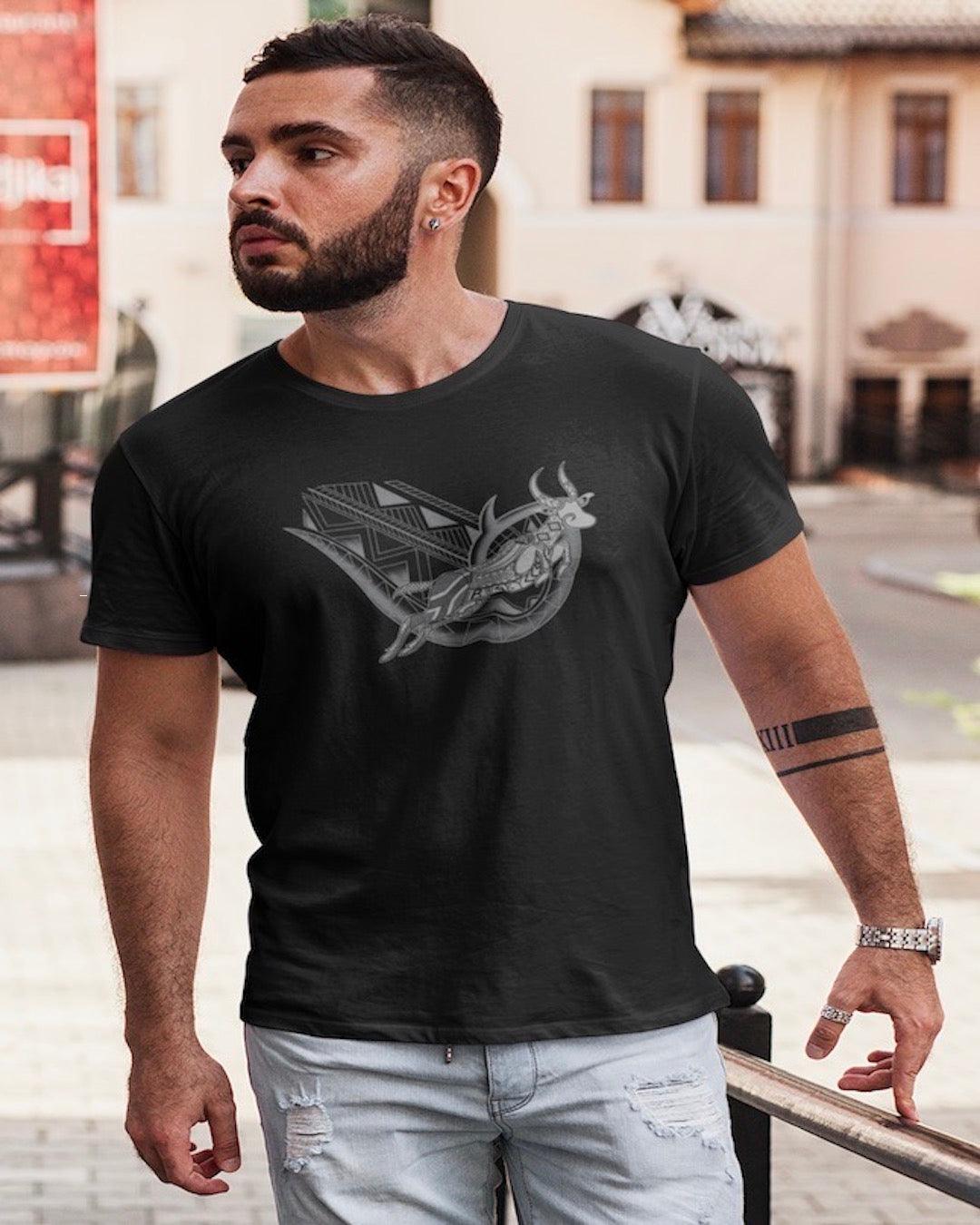 RUGBYNOMAD black organic cotton classic heavyweight streetwear t shirt with a south africa springbok artistic design on a male model with dark hair, dark beard, arm tattoos, wearing light colored blue jeans walking down the street, bright colored buildings behind him