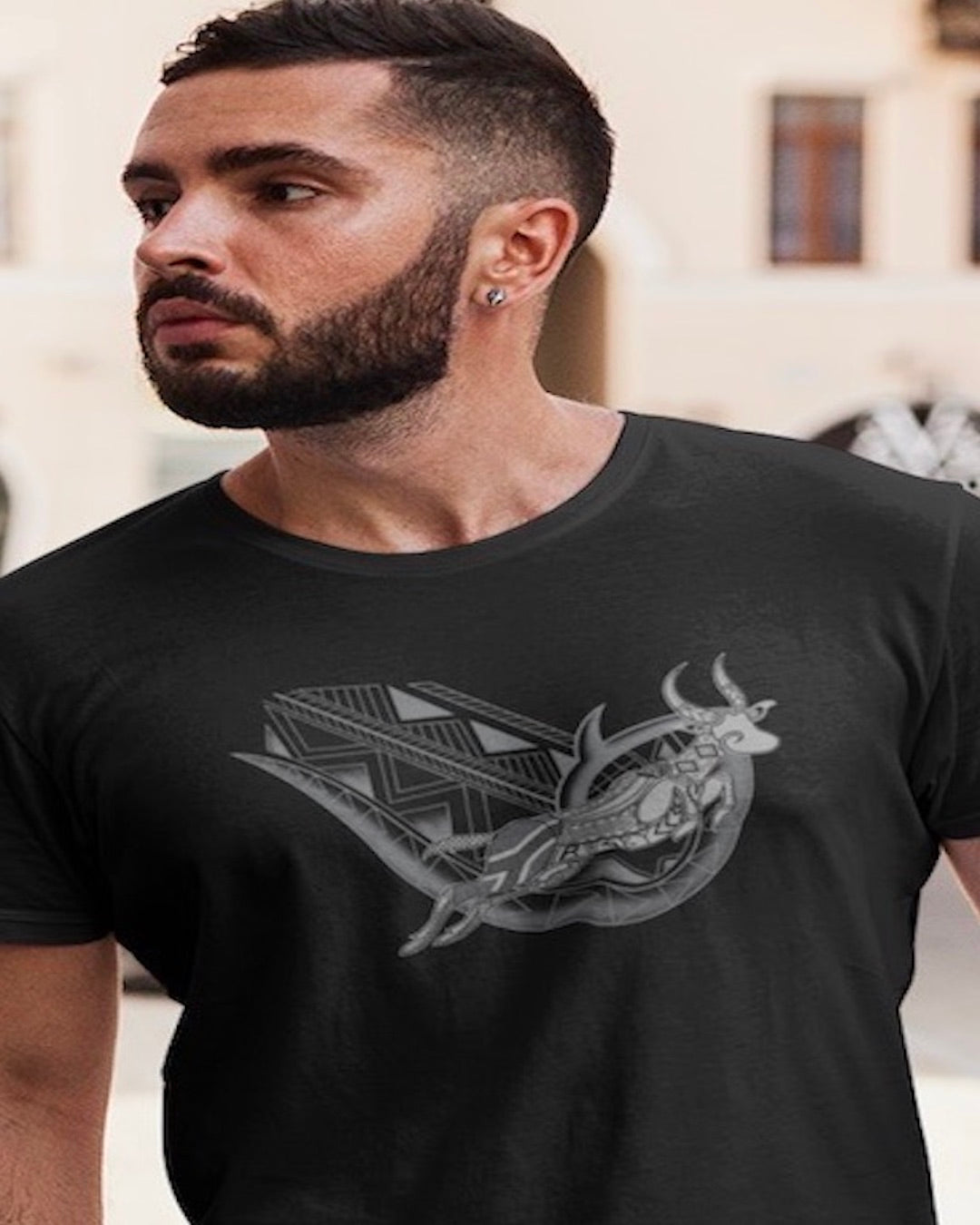 RUGBYNOMAD black organic cotton classic heavyweight streetwear t shirt with a south africa springbok artistic design on a male model with dark hair, dark beard, arm tattoos, wearing light colored blue jeans walking down the street, bright colored buildings behind him