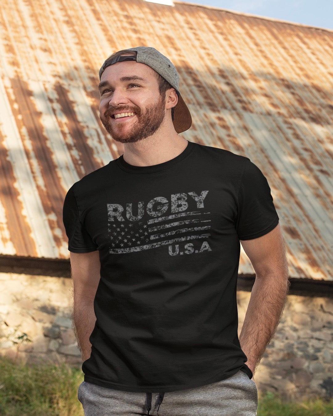 RUGBYNOMAD black organic cotton classic heavyweight streetwear t shirt with a usa flag and rugby artistic design on a male model with dark hair and beard wearing a gray baseball cap backwards with hands in his pockets standing in front of a rusty barn roof on a farm looking to his right