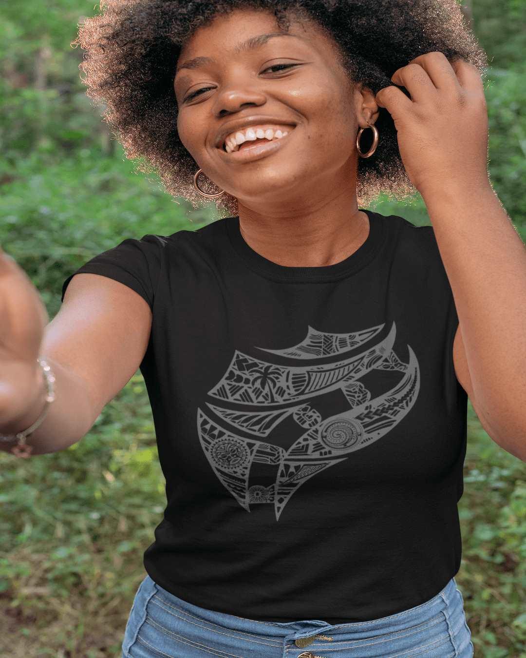 RUGBYNOMAD blue sage color organic cotton women's streetwear t shirt on a female model standing in front of a green jungle with one hand in her dark hair smiling with fiji artistic tribal design