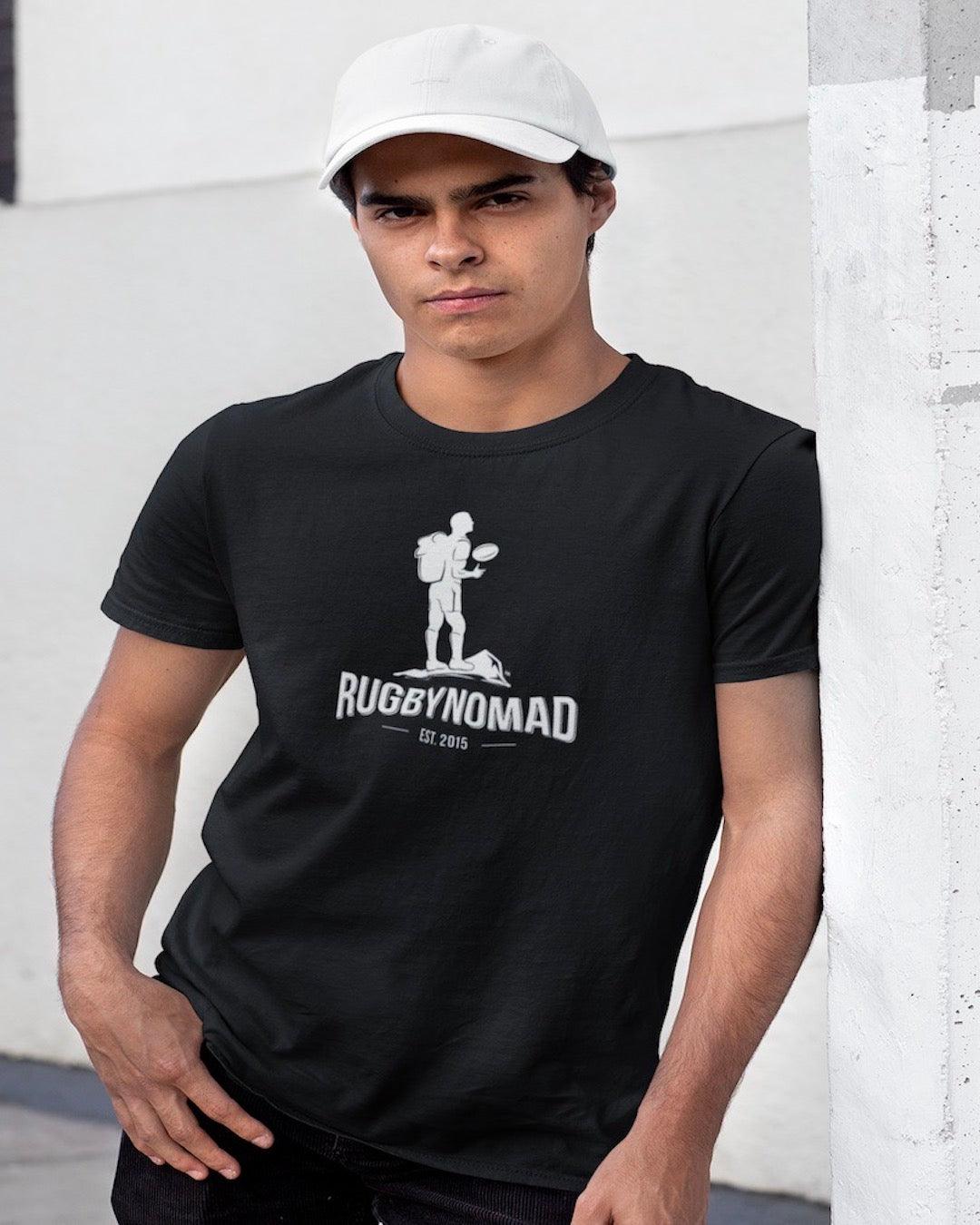 RUGBYNOMAD black organic cotton heavyweight streetwear t shirt with RUGBYNOMAD original complete logo in white  est 2015 on a male model leaning against a concrete wall wearing a white hat with his hands in his pockets, dark hair, with a tough look on his face